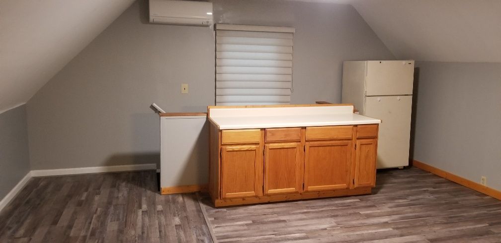 Kitchenette with wooden cabinets, refrigerator, and window. Gray walls and flooring, angled ceiling.