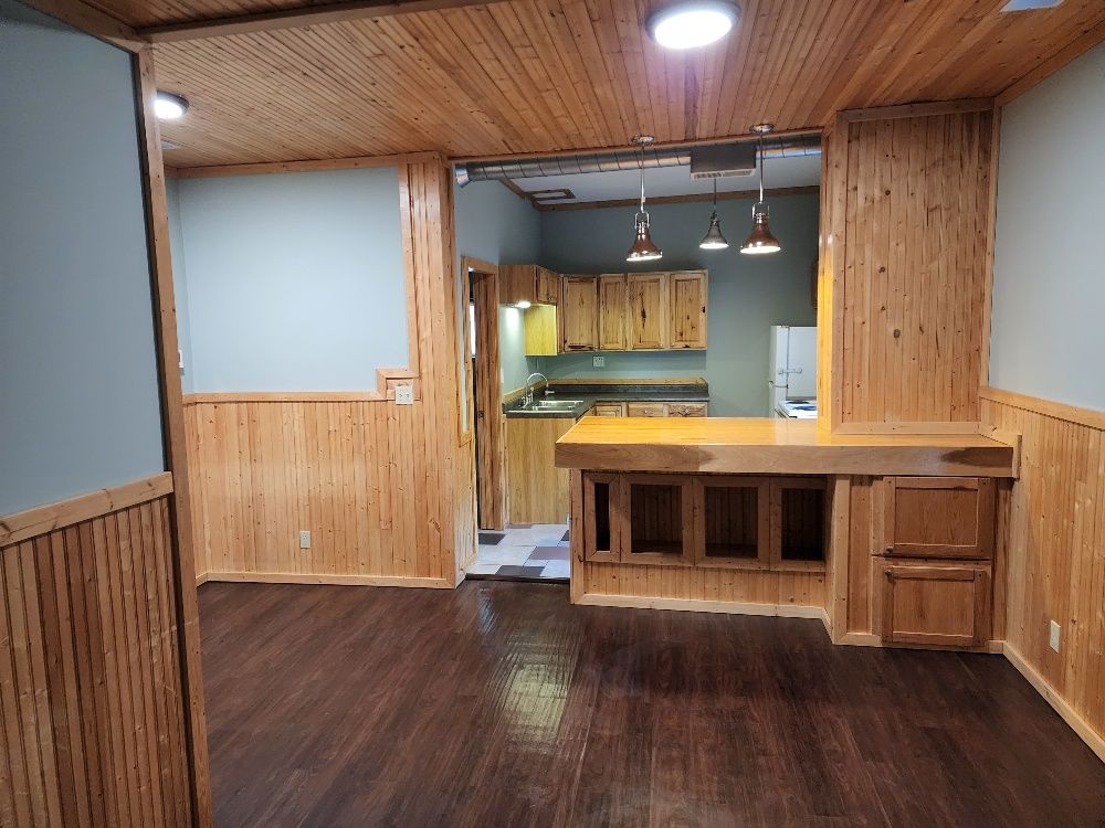 Interior view of a room with wood paneling, dark wood floor, and a kitchen area visible.