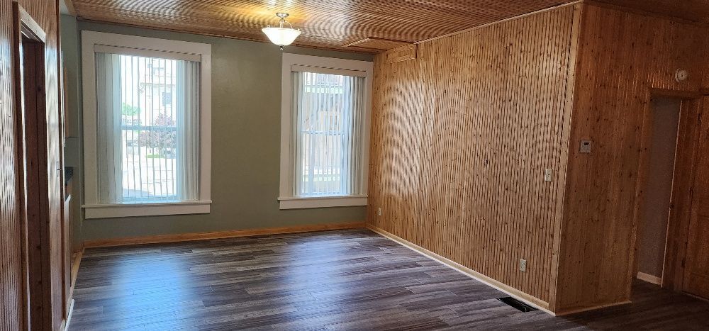 Interior view of a room with wooden paneling on walls and ceiling, two windows, and dark flooring.