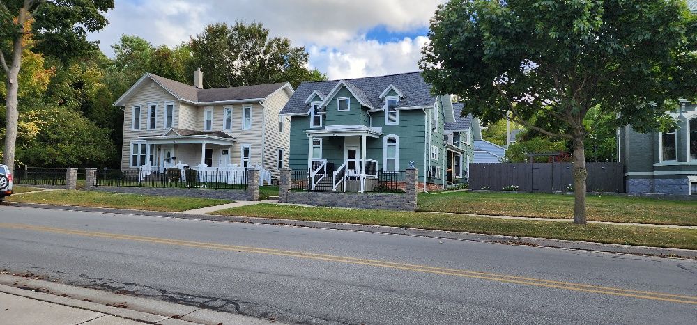 Houses along a street. Light-colored house on the left, a teal-colored house in the middle and green house on the right, trees and sky in the background.