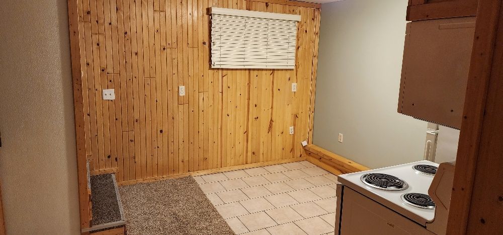Interior of a small room. Wood-paneled walls, tile floor, stove, and a window with a blind are visible.