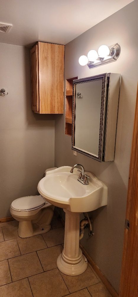 Bathroom with pedestal sink, toilet, wall cabinet, and mirror with light fixture.