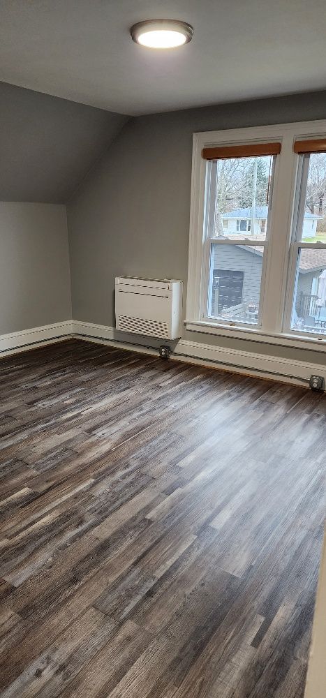 Empty room with grey walls, dark wood-look floor, window with brown trim, and a ceiling light.