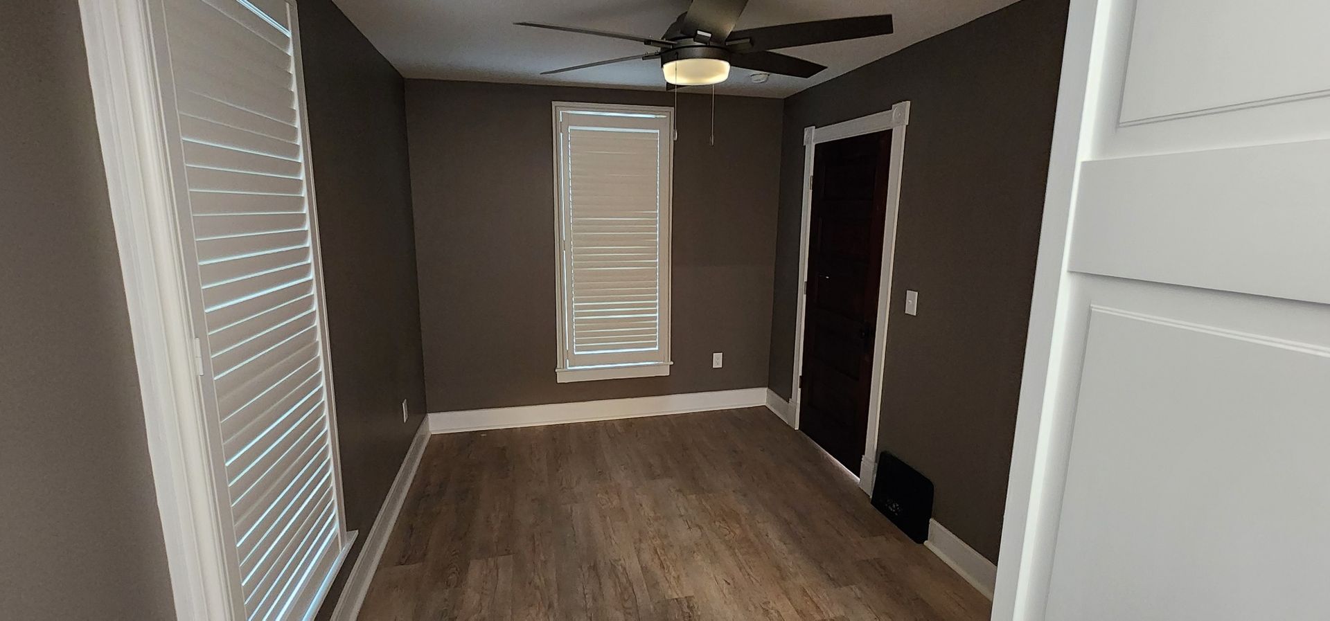 Empty room with brown walls and hardwood floors; window with blinds, ceiling fan, and closed door.