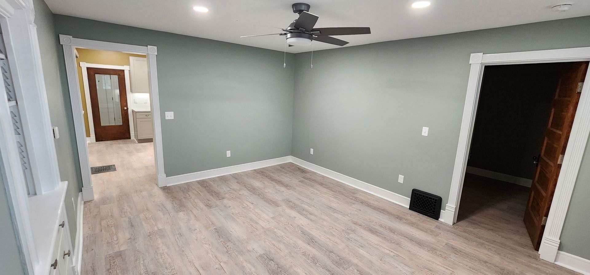 Empty room with grey-green walls, light wood-look floors, and three doorways. A ceiling fan is visible.