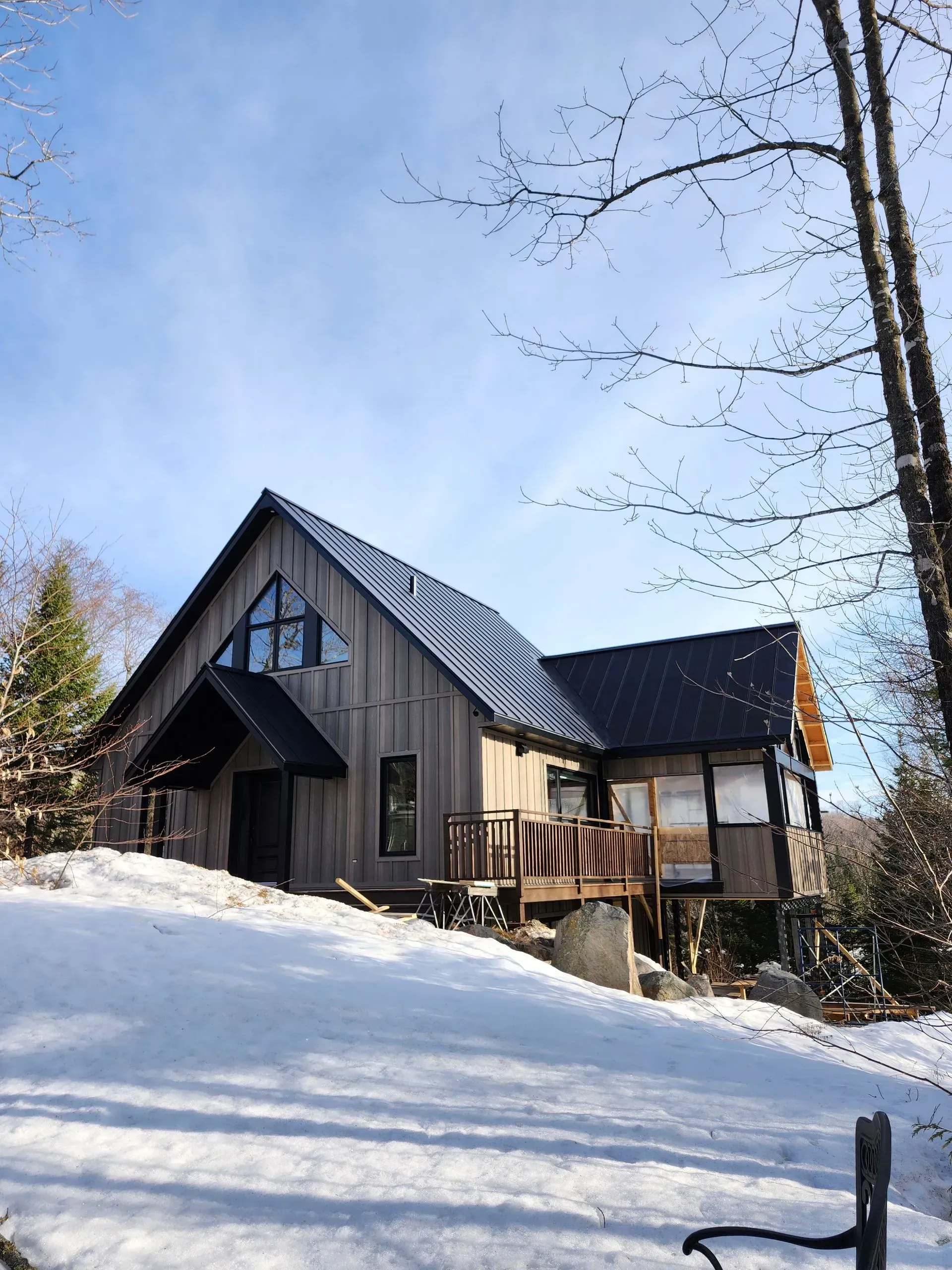Cabane enneigée au toit de métal sombre, sur un versant ensoleillé en hiver, entourée d'arbres dénudés.
