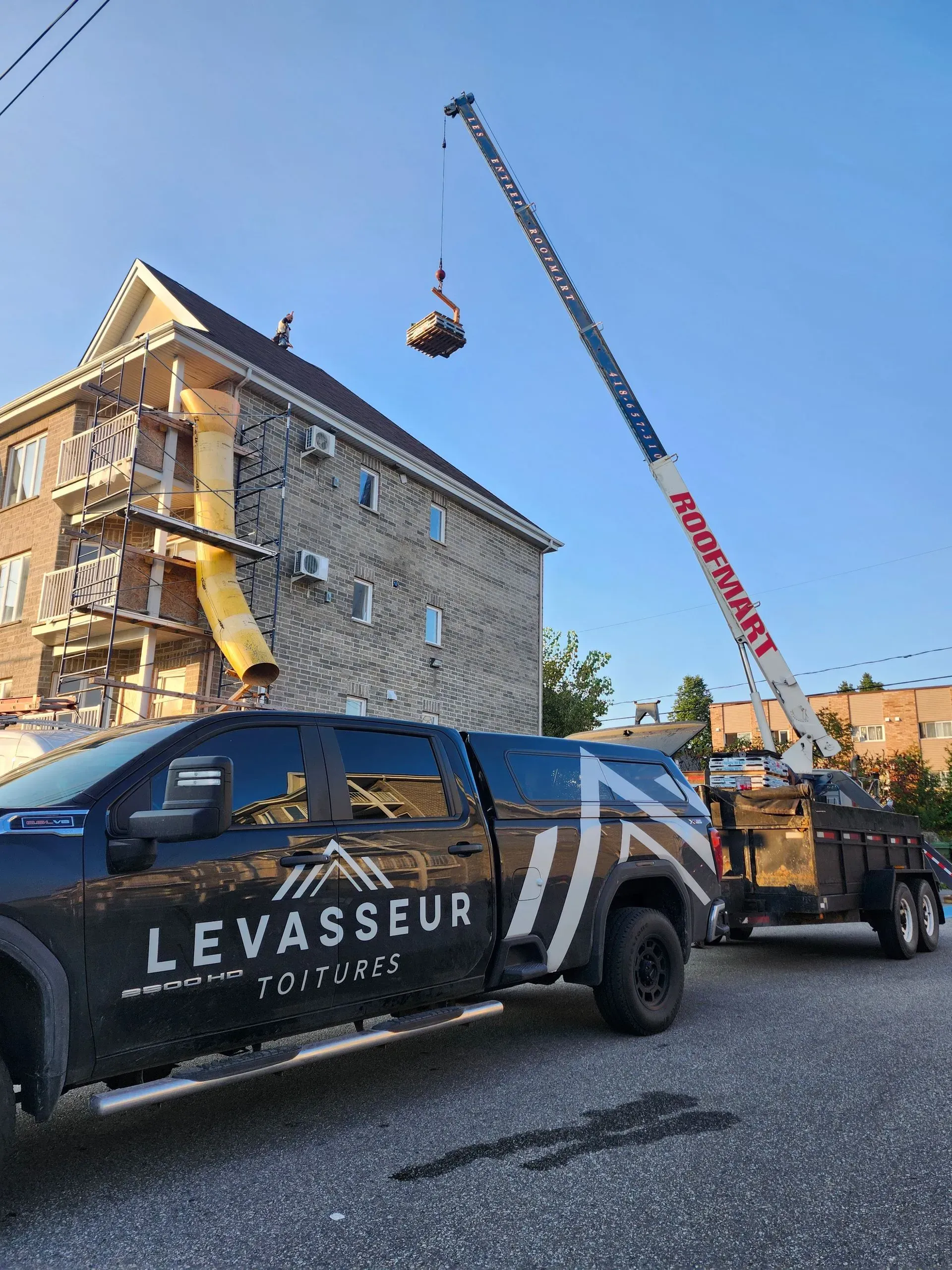 Une grue soulève un paquet près d'un bâtiment en pierre, à côté d'un camion Vasseur, dans une rue ensoleillée.