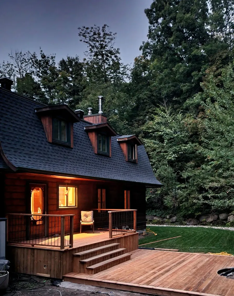 Chalet confortable en bois sombre avec véranda et terrasse éclairées au crépuscule, entouré d'arbres