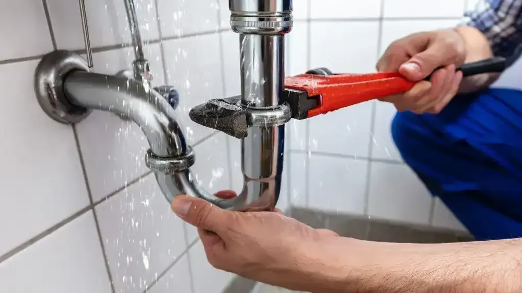 Plumber using a red wrench to tighten a bathroom sink pipe under a tiled wall.