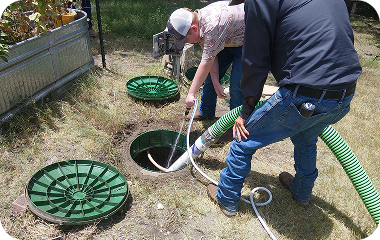Two workers using hoses to inspect a manhole in a yard, with green sewer covers nearby.