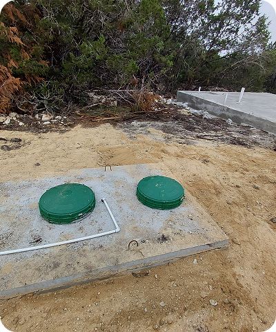 Two green circular utility covers on a concrete pad in a sandy, brushy area