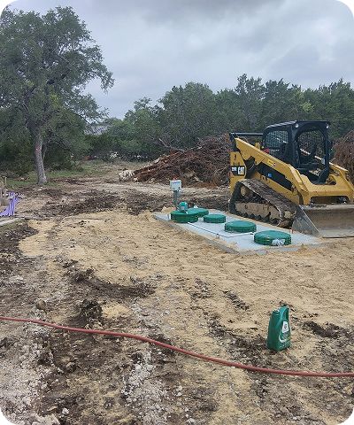 Construction site with a skid-steer, dirt piles, and green utility covers near trees