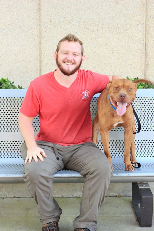 Man in red shirt sitting on a bench beside a brown dog with tongue out