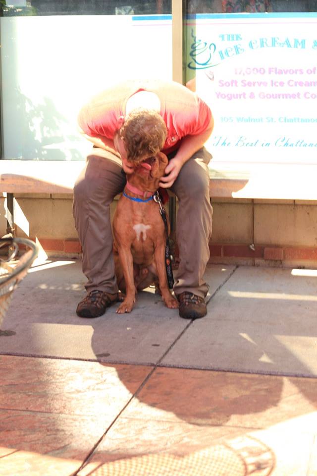 Person crouches to hug a brown dog on a sidewalk outside a storefront.