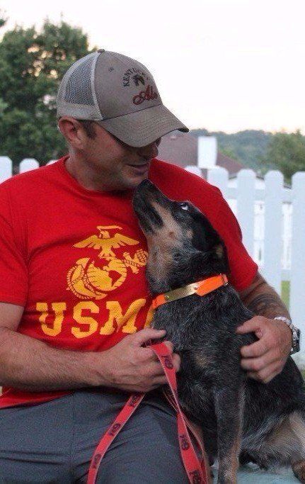 Man in red shirt and cap holds a dog on a red leash, looking at each other outdoors.