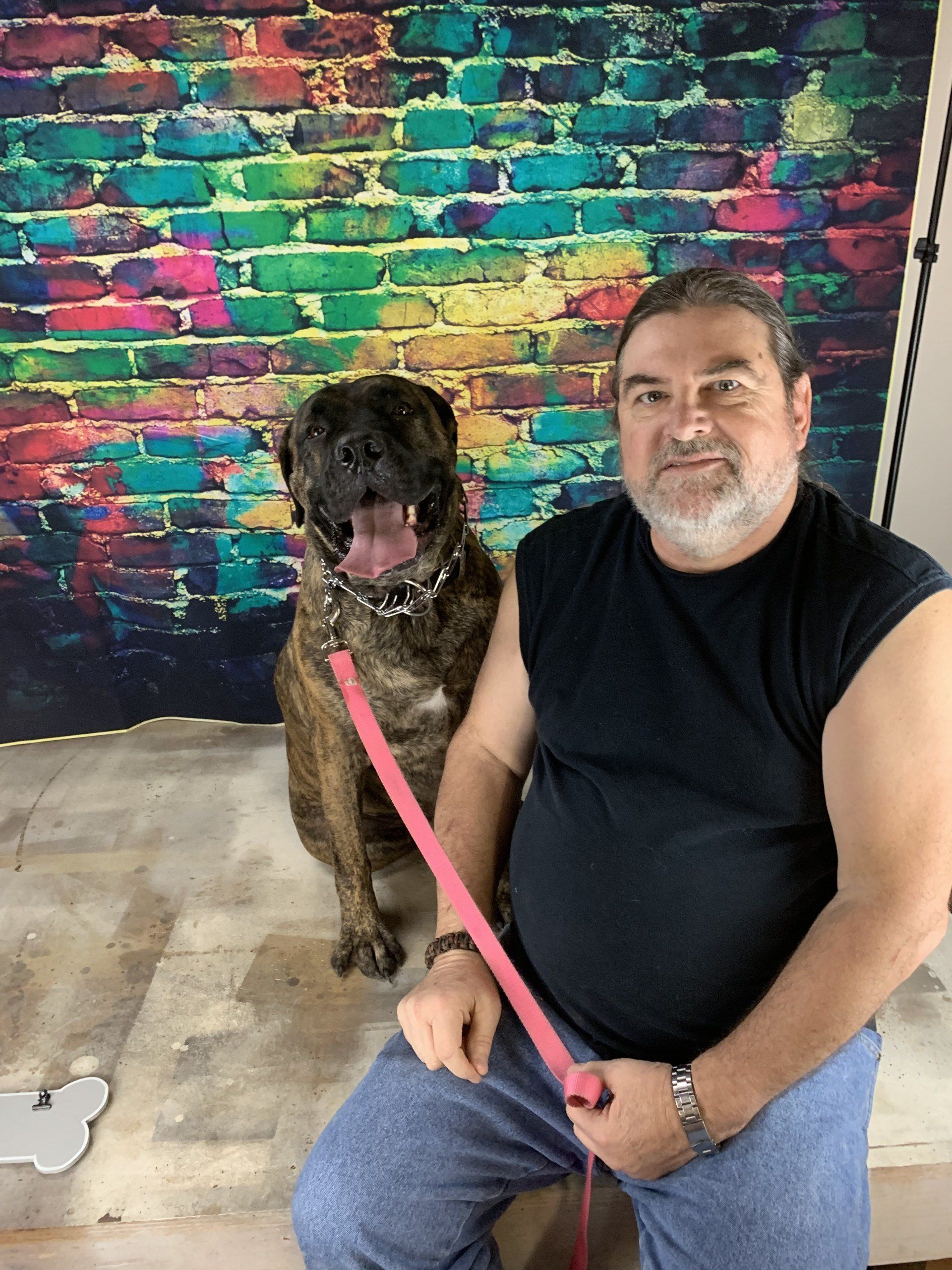 Man seated beside a brown dog on a pink leash in front of a colorful brick wall
