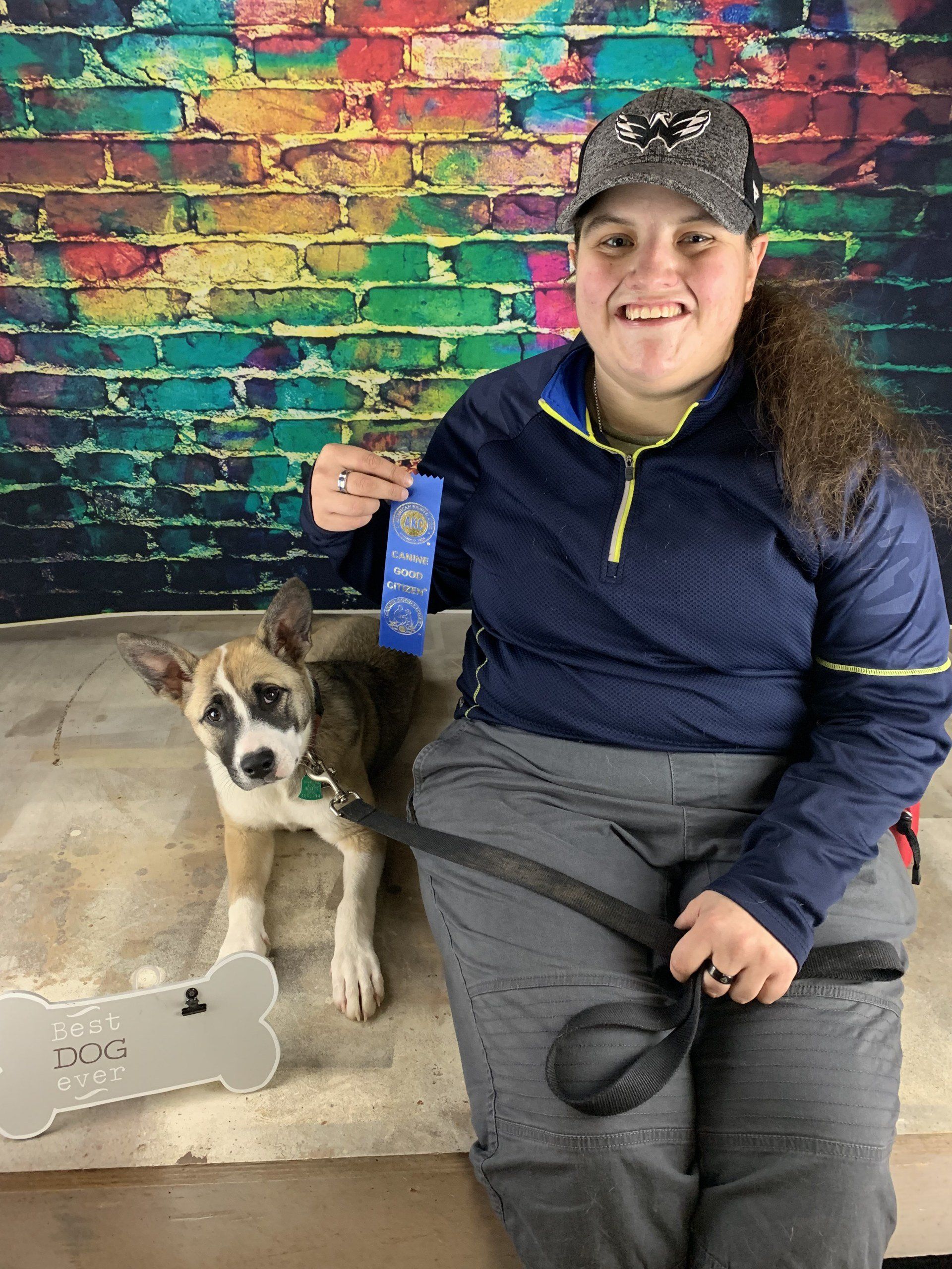 Person sitting beside a dog, holding a blue ribbon in front of a colorful brick wall.