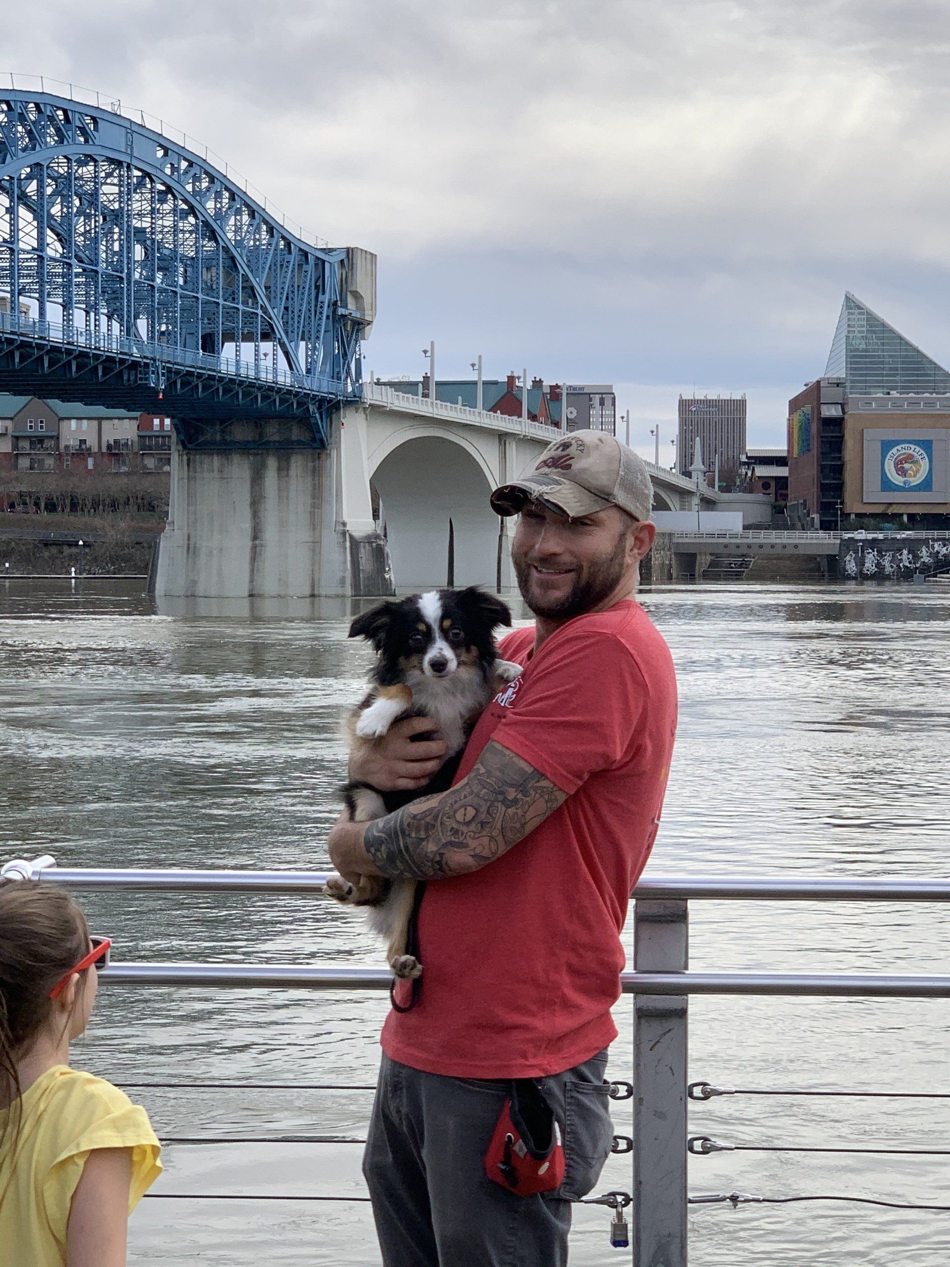 Man holding a small black-and-white dog on a riverside walkway with a blue bridge in the background