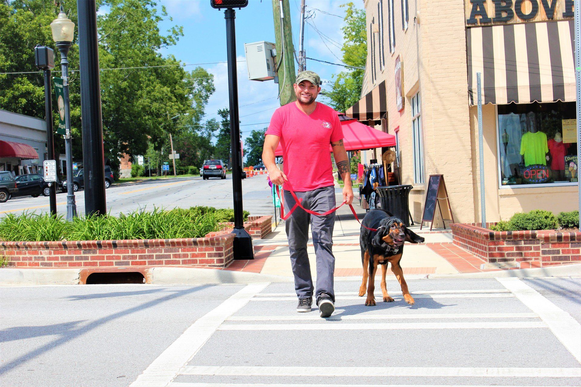 Man in a bright pink shirt walking a dog across a city street on a sunny day