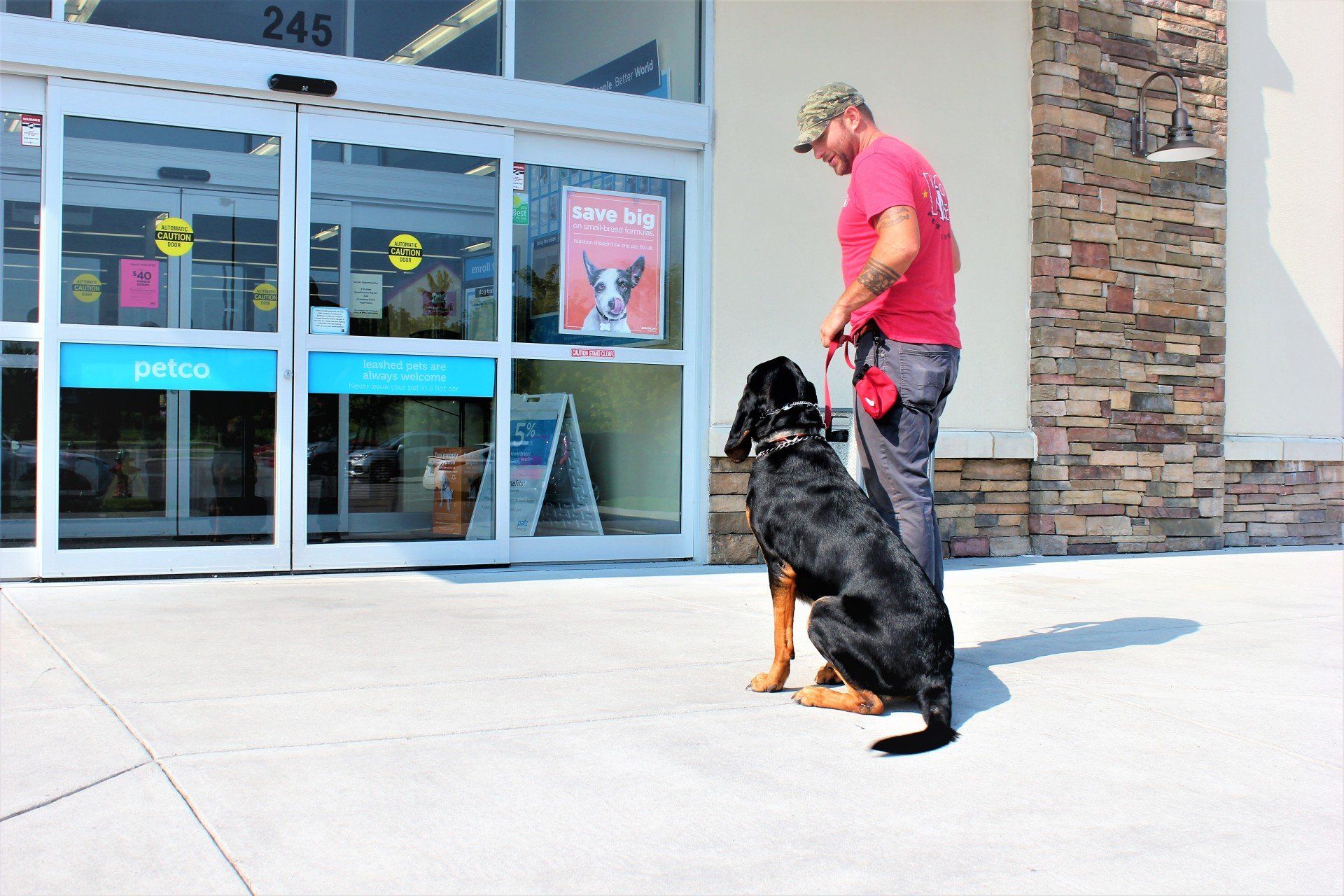 Person in a red shirt walking a large black dog outside a store entrance.