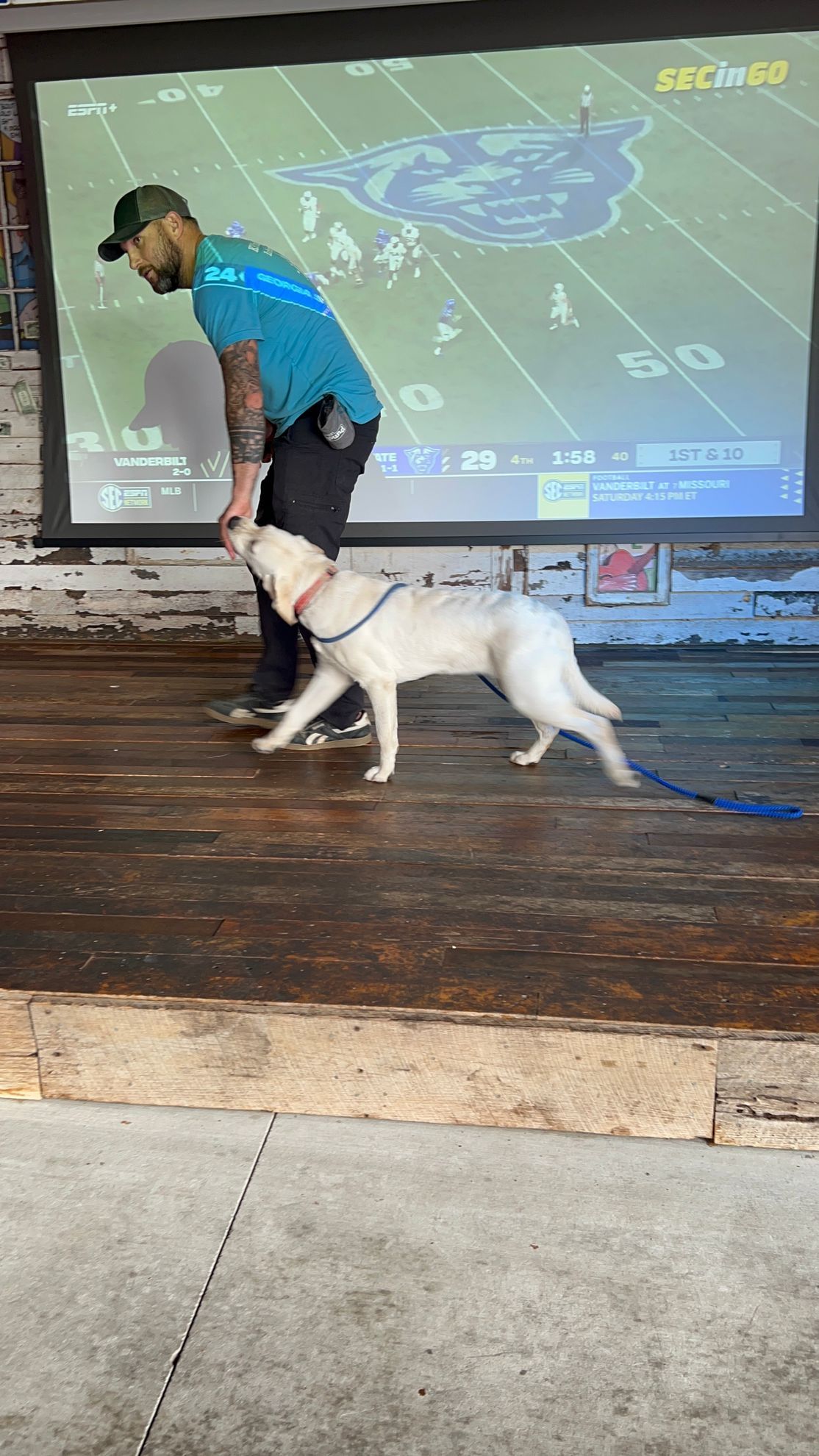 Man in teal shirt with a white dog on a leash in front of a projected screen on a wooden stage