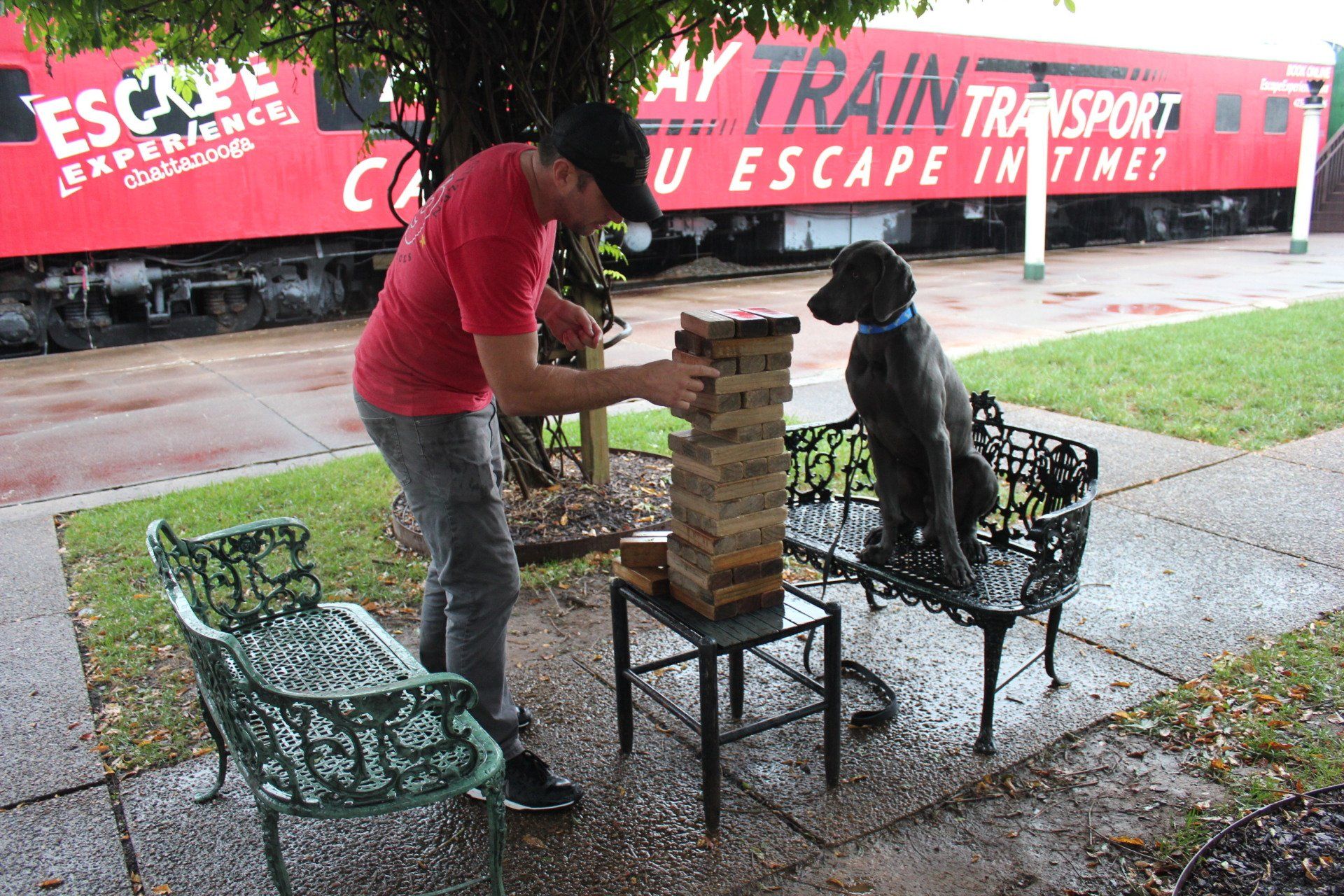 Two people play giant Jenga at a park table near a red “Escape” train display