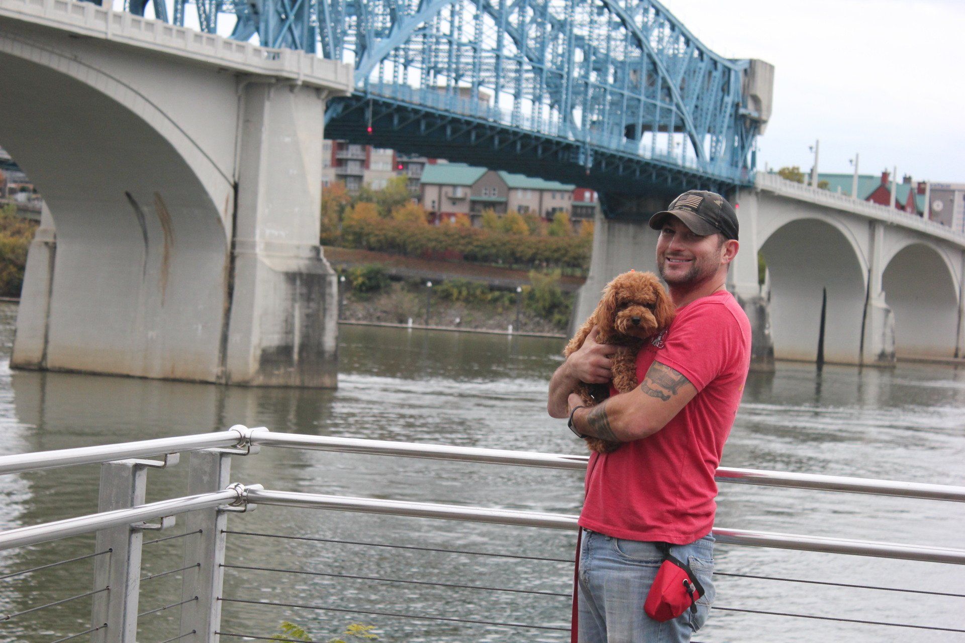 Man holding a small brown dog by a river near a blue bridge, wearing a red shirt and cap.