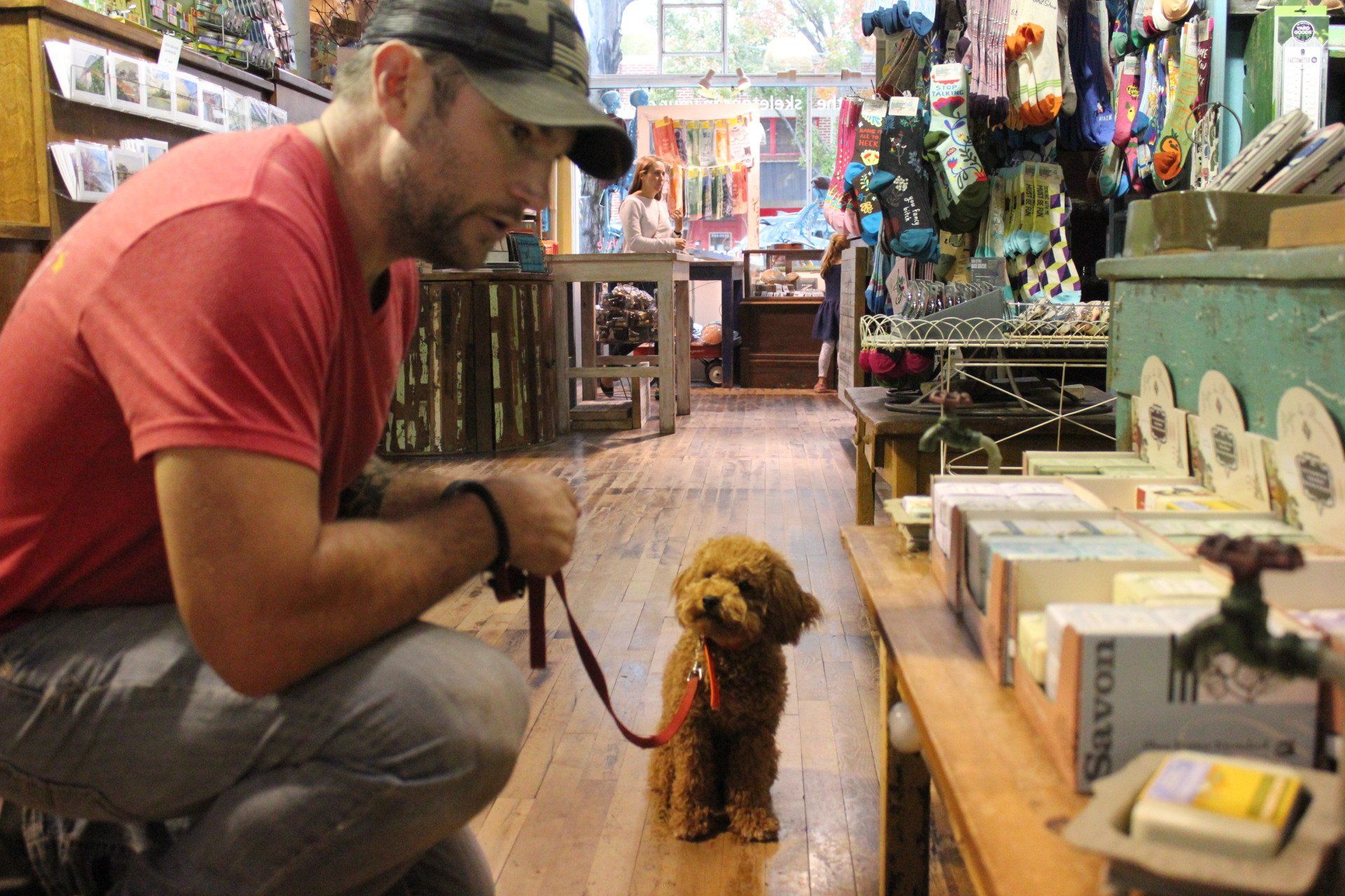 Man kneels in a shop aisle, holding a leash to a small dog facing him.