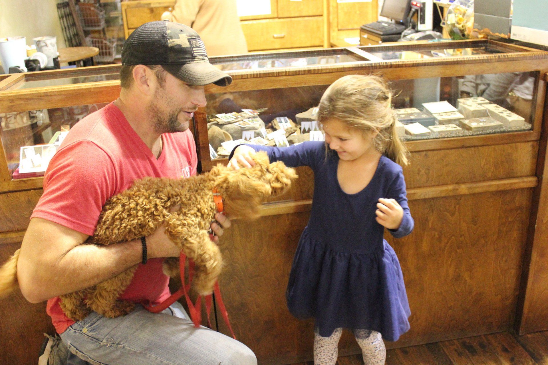 Man in red shirt holding a lamb while a child in blue dress pets it at a farm counter