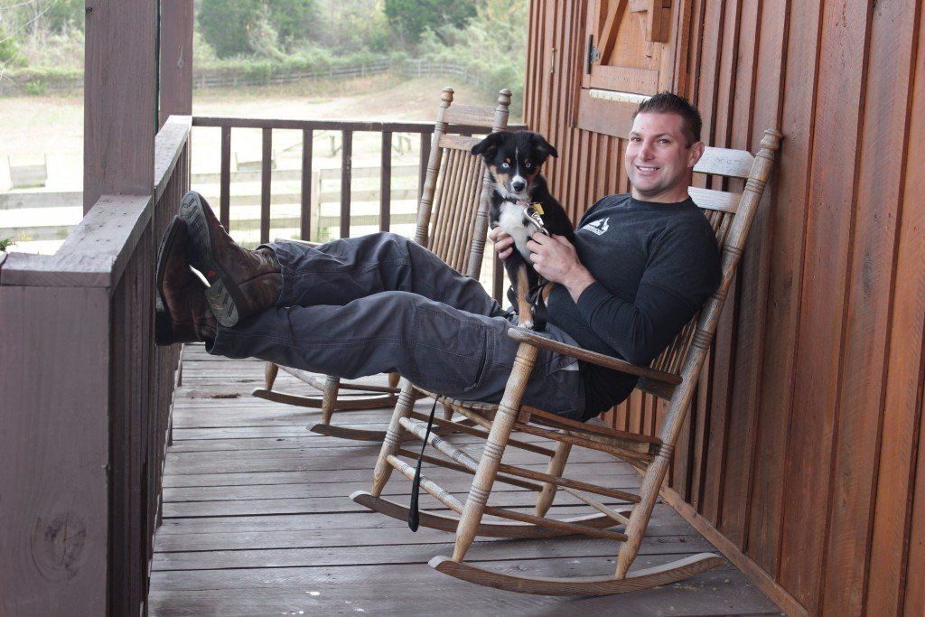 Man reclining on a wooden porch rocking chair, holding a black-and-white dog.