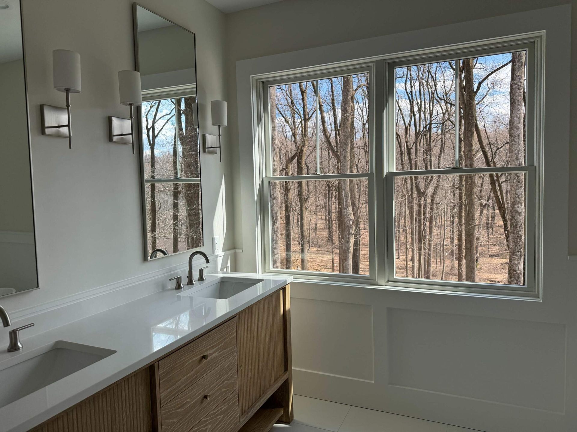 Bathroom with a double sink vanity, mirrors, and a large window overlooking trees.