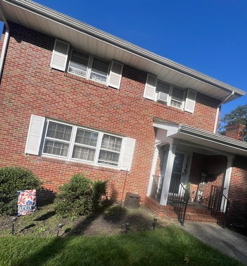 Red brick two-story house with white shutters. Green lawn, blue sky, and a small porch are visible.