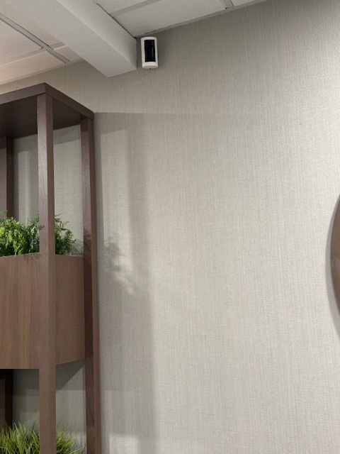 Security camera mounted on a beige wall in an office setting. A plant shelf is in the left corner.