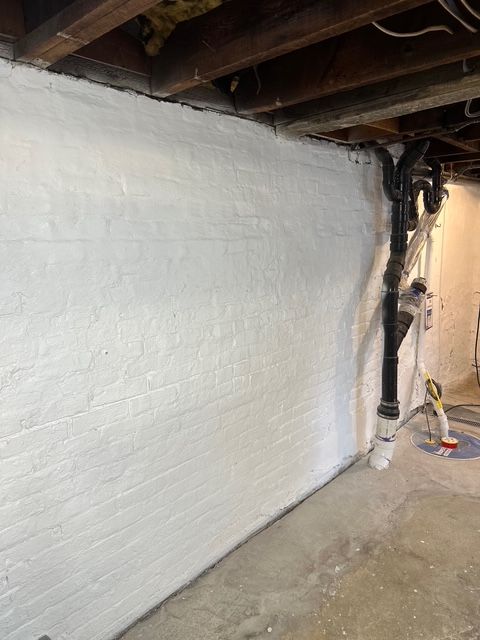 White-painted brick wall in a basement, next to pipes and ceiling beams. Concrete floor.