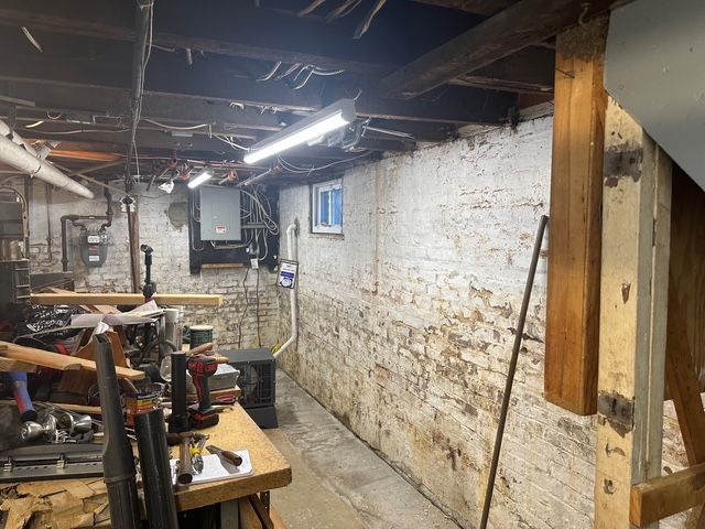 Basement workshop with a brick wall, cluttered workbench, and exposed pipes and beams.
