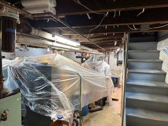 Basement interior with plastic-covered equipment, exposed pipes, wooden beams, and stairs leading up.