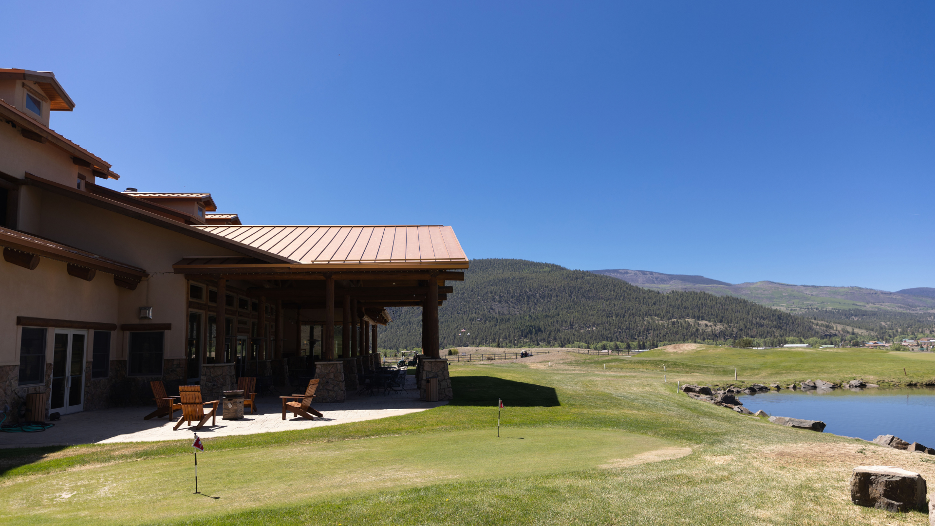 Mountain lodge patio beside a grassy lakeside under a clear blue sky at Rio Grande RV Resort
