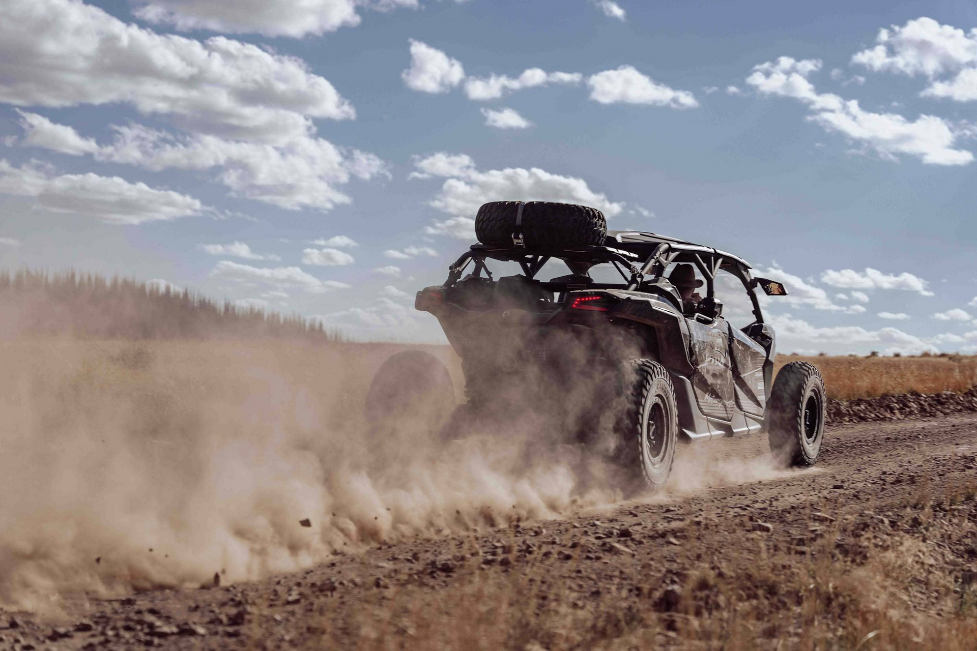Black off-road vehicle driving on dirt road, kicking up dust under a cloudy blue sky.