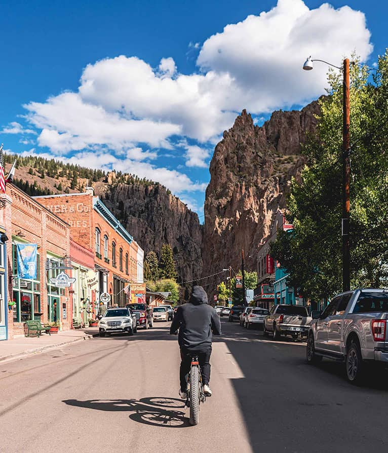 A cyclist rides down a street in a mountain town, flanked by buildings and tall rock cliffs under a blue sky.