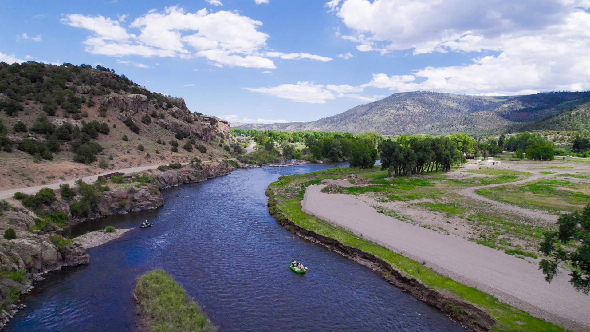 Winding blue river through green valley with sandy banks, trees, and hills under a partly cloudy sky in Rio Grande RV Resort