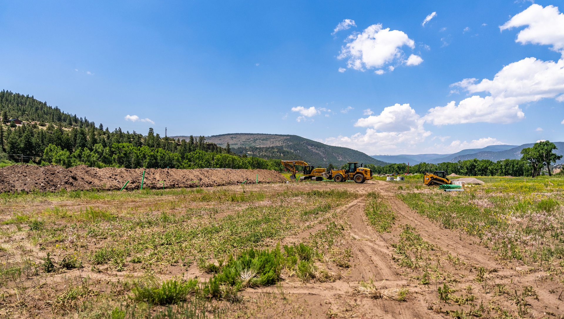 Wide dirt field with tire tracks, green hills, and scattered clouds under a bright blue sky.