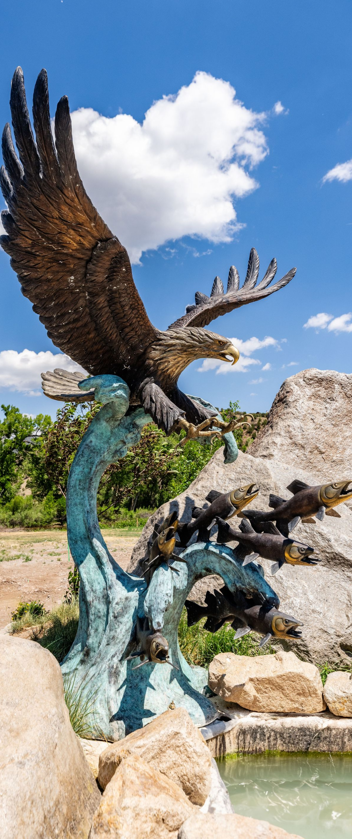 Bronze eagle sculpture with outstretched wings beside a turquoise pool under a blue sky with clouds
