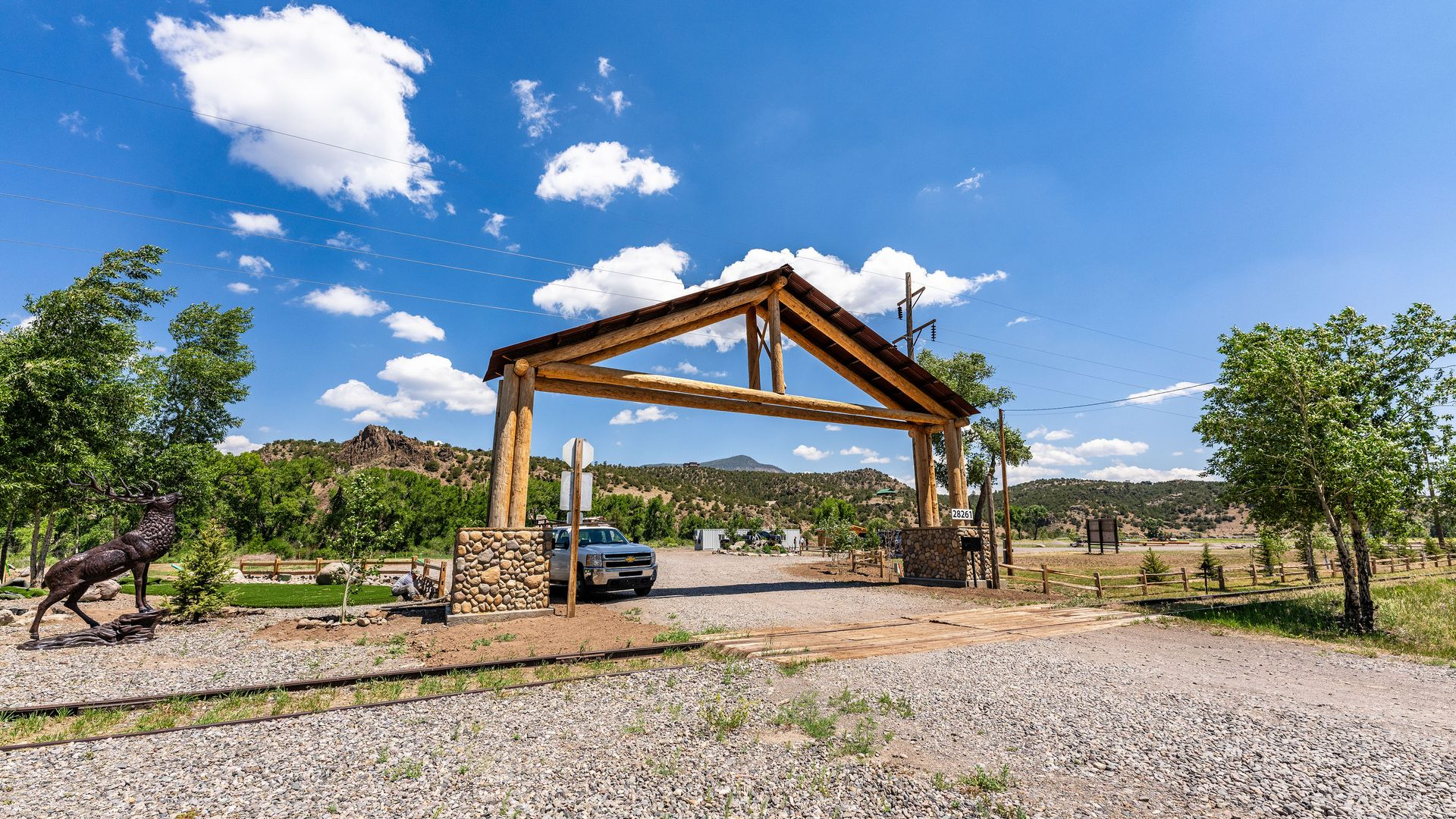 Wooden pavilion under construction on a gravel lot, with trees and hills under a blue sky