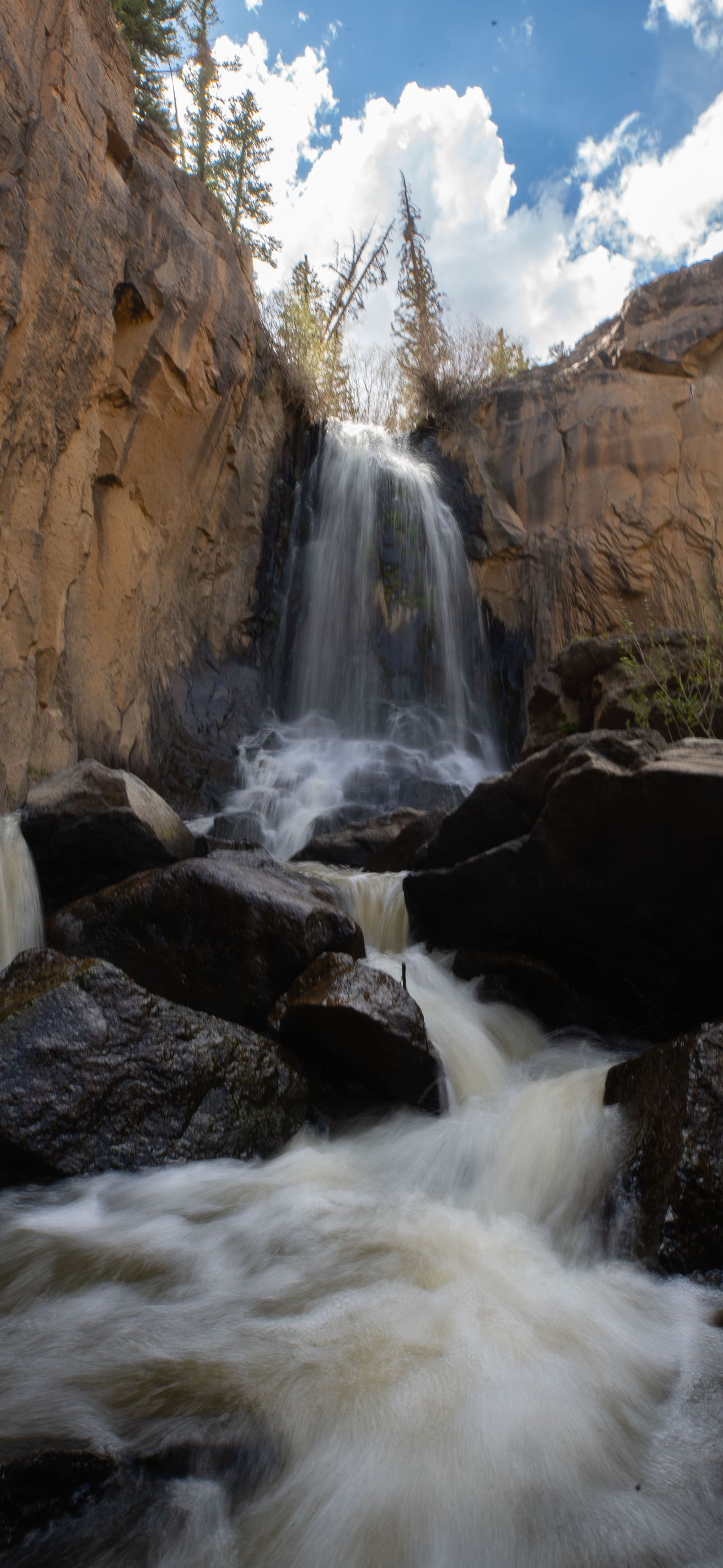 Waterfall cascading through a rocky canyon with white water and pine trees under a cloudy sky