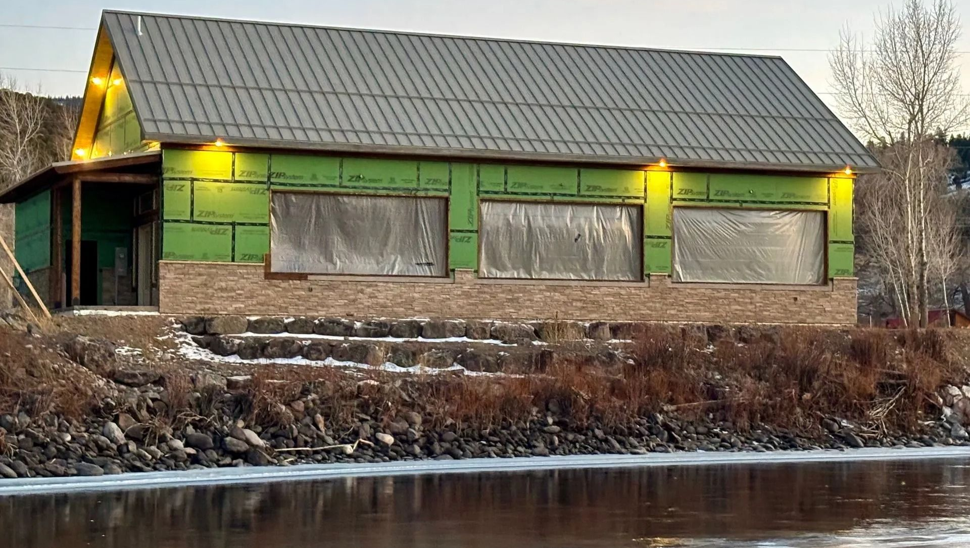 Rustic green building by a river at dusk, with lit exterior lights and a snowy roof.