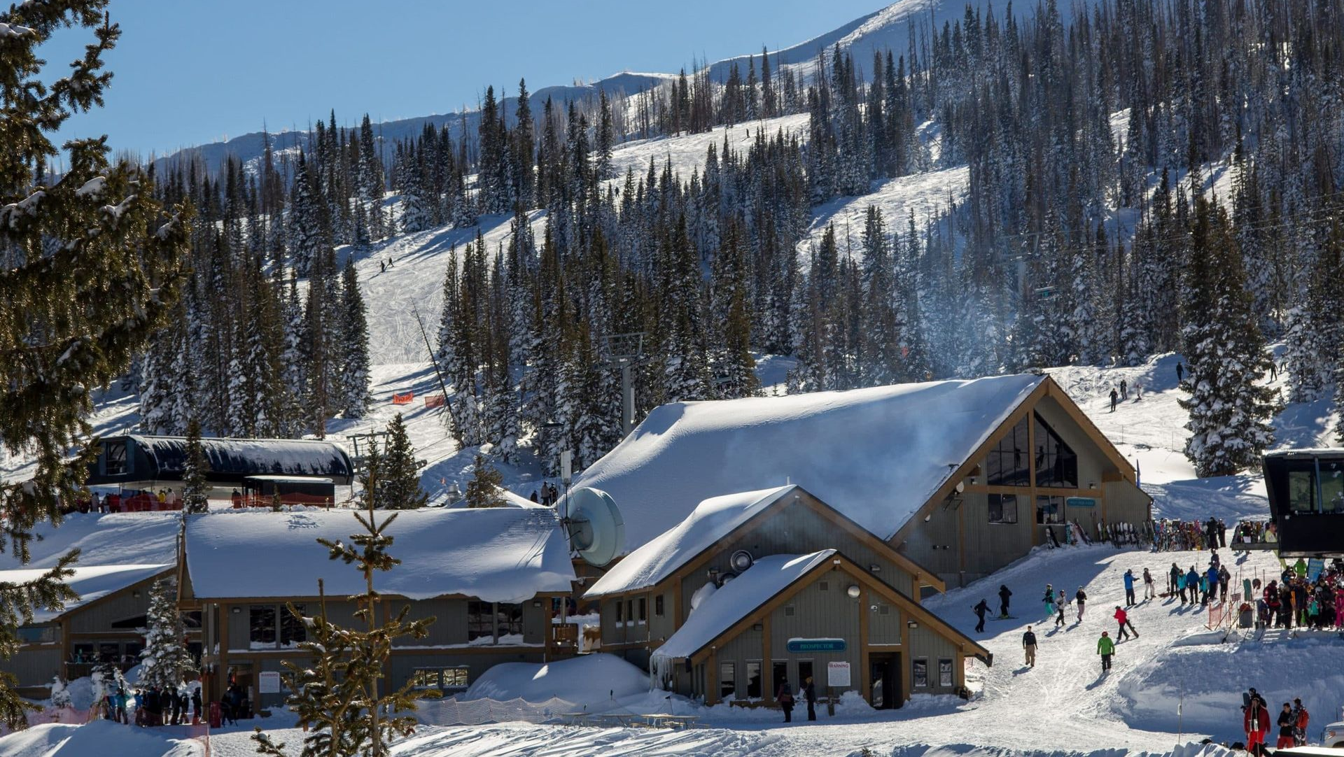 Snowy mountain village with chalets, pine trees, and skiers under a clear blue sky.