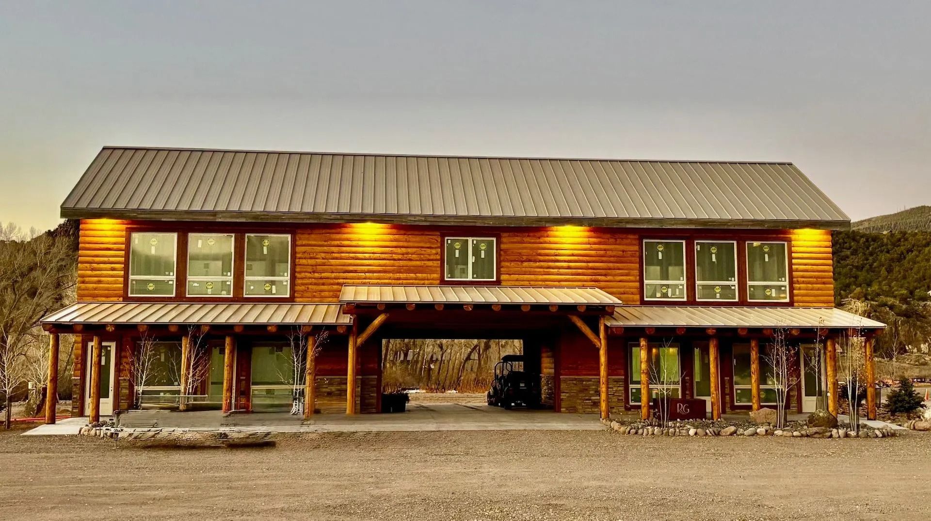 Rustic two-story wooden house with a wraparound porch and a large open carport, lit at dusk.