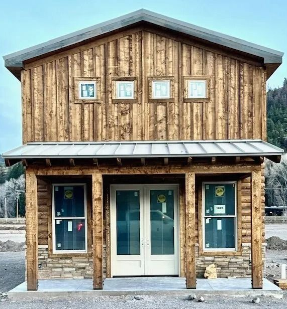 Rustic two-story wooden building with glass double doors and windows on a gravel lot, mountains behind.