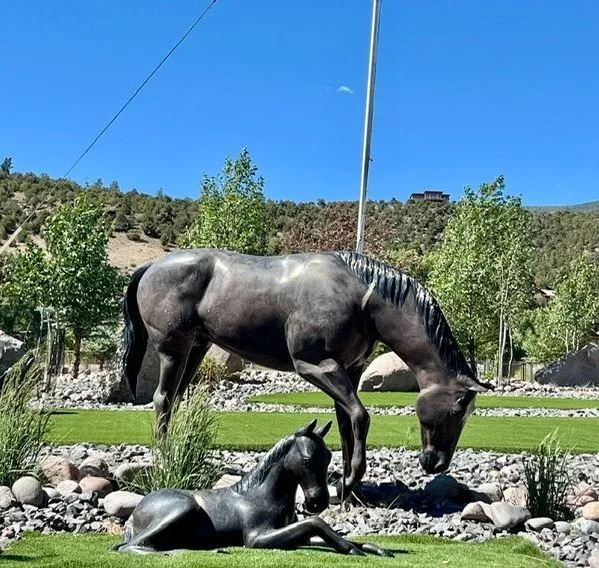 Two dark horse statues in a sunny garden, one standing and one lying down, with trees and hills behind them.