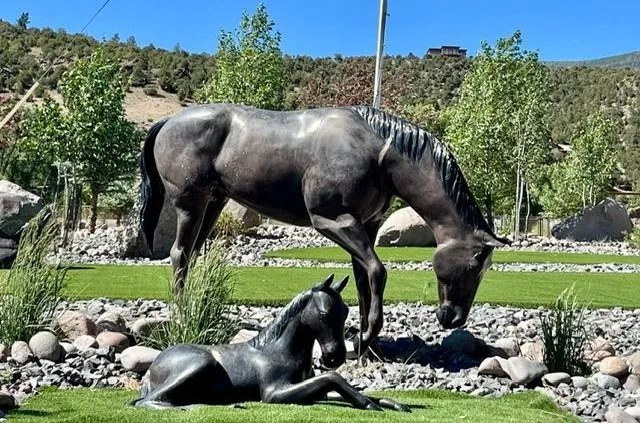 Bronze horse statues in a landscaped garden, one standing and one resting on grass.